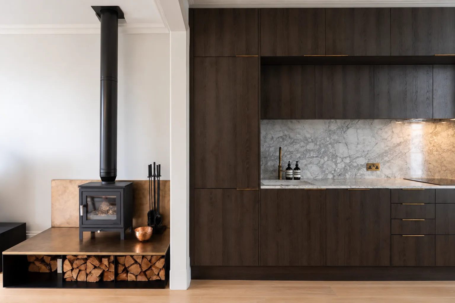 Open-plan kitchen and living space in Chelsea: a log burner on a bronze-topped plinth with a log store, beside a dark oak kitchen with a marble splashback
