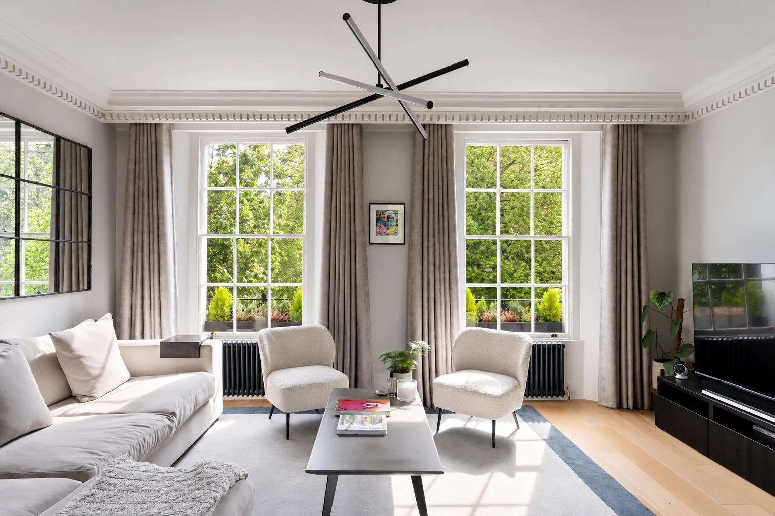 Living room in a Chelsea period home with two tall sash windows, full-height curtains, dentil cornicing, a pale sofa and two armchairs