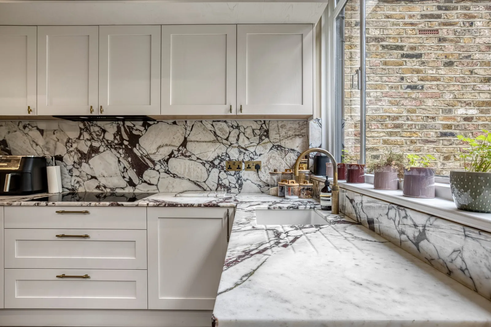 Shaker kitchen with marble worktops and splashback, brass tap and a window onto the side return