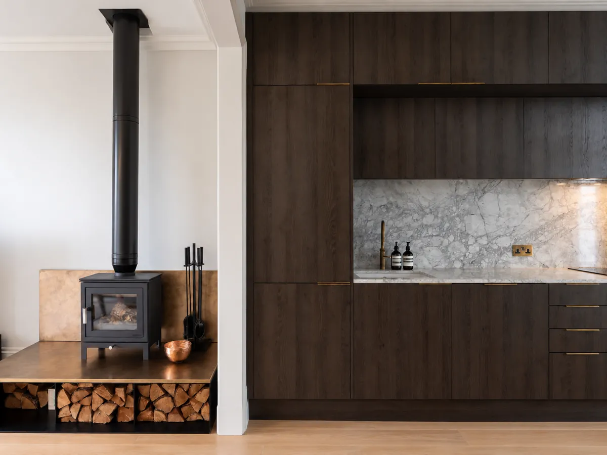 Open-plan kitchen and living space in Chelsea with a log burner on a bronze-topped plinth beside a dark oak kitchen and marble splashback